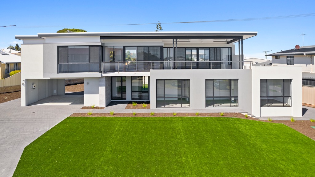 Modern two-storey house with large windows, flat roof, balcony, and manicured front lawn