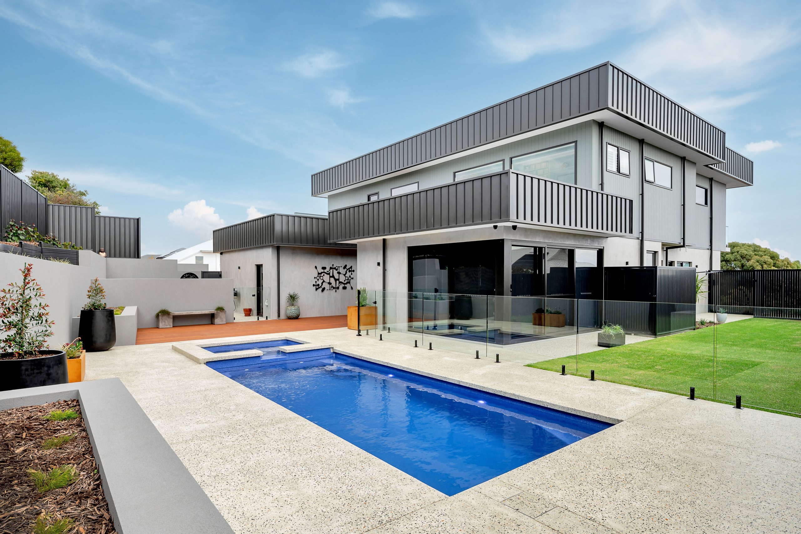 Modern double-storey home with a custom swimming pool, raised spa, glass pool fencing and concrete entertaining area, set against a manicured lawn