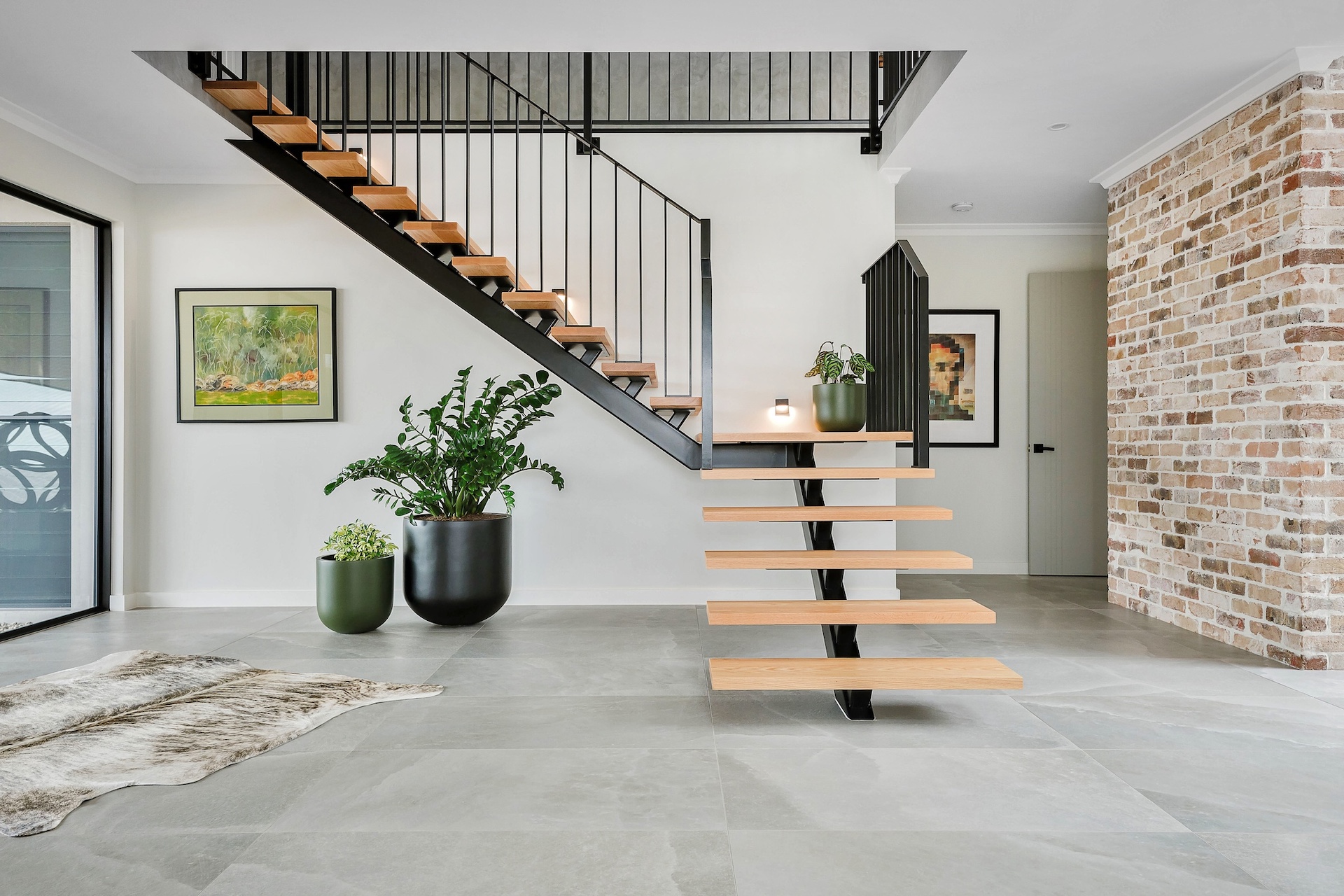 Open-plan entryway featuring a bespoke floating timber and steel staircase, exposed recycled brick feature wall and large-format tiled flooring