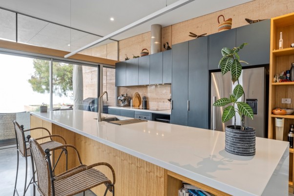 Modern open-plan kitchen with a long stone island bench, timber cabinetry and a rammed-earth feature wall, looking out to a covered alfresco area.