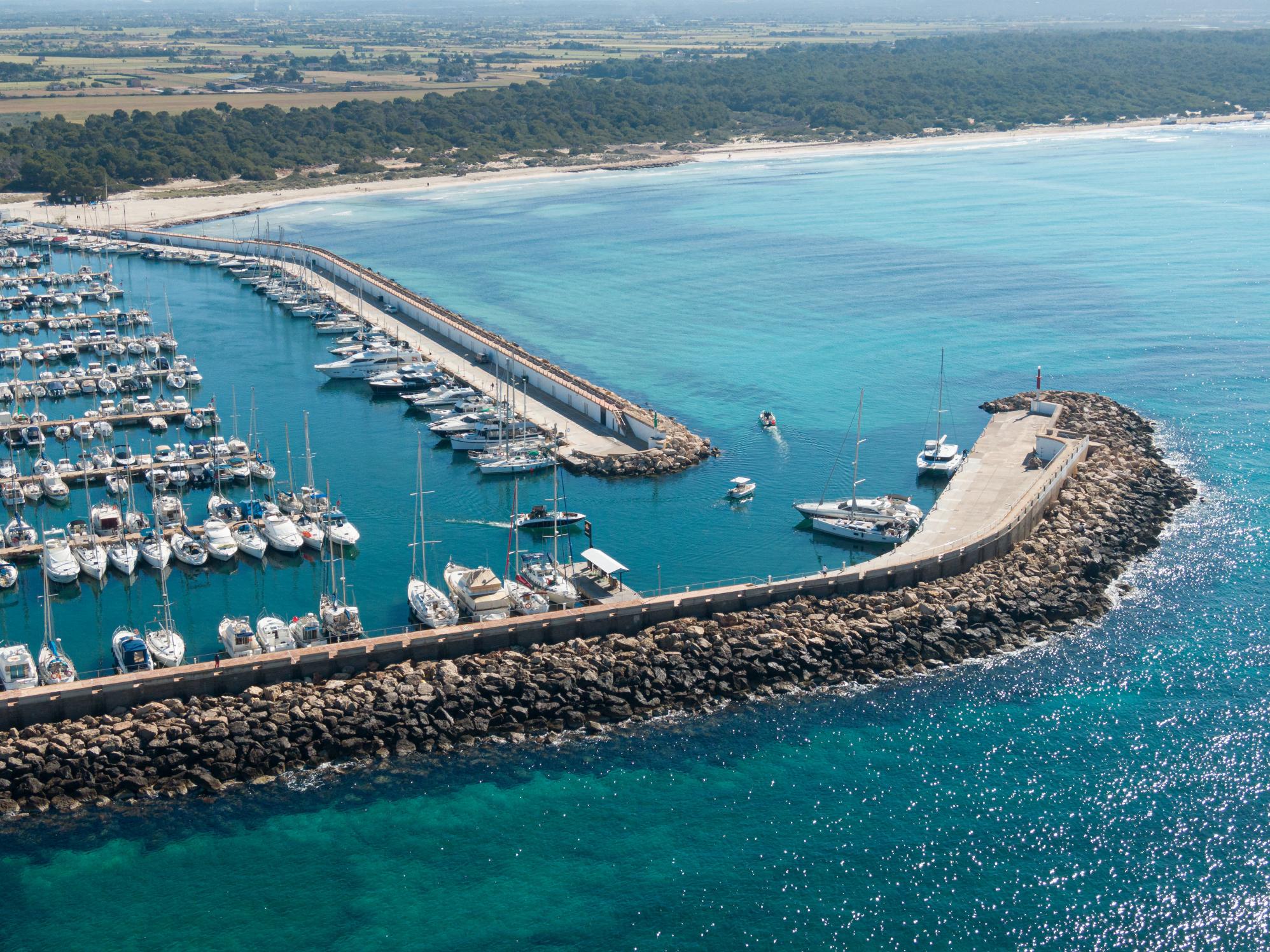 A coastal marina with a number of moored boats, a curved stone breakwater, turquoise water and a sandy beach