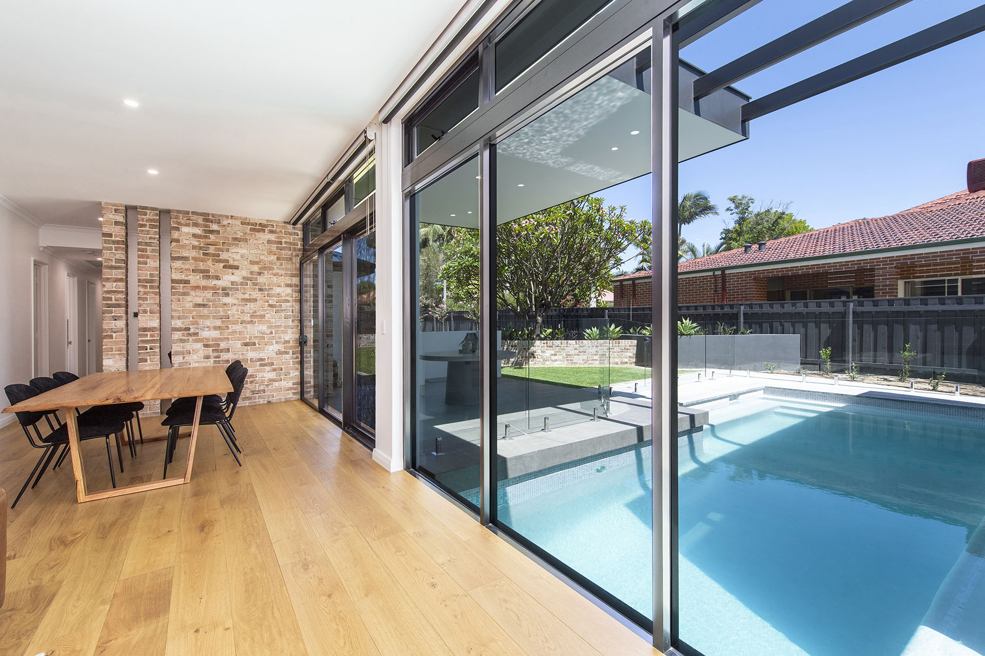 A dining room with wooden plank floorboards and a mortar brick feature wall with glass doors leading to the backyard with a pool and grass area